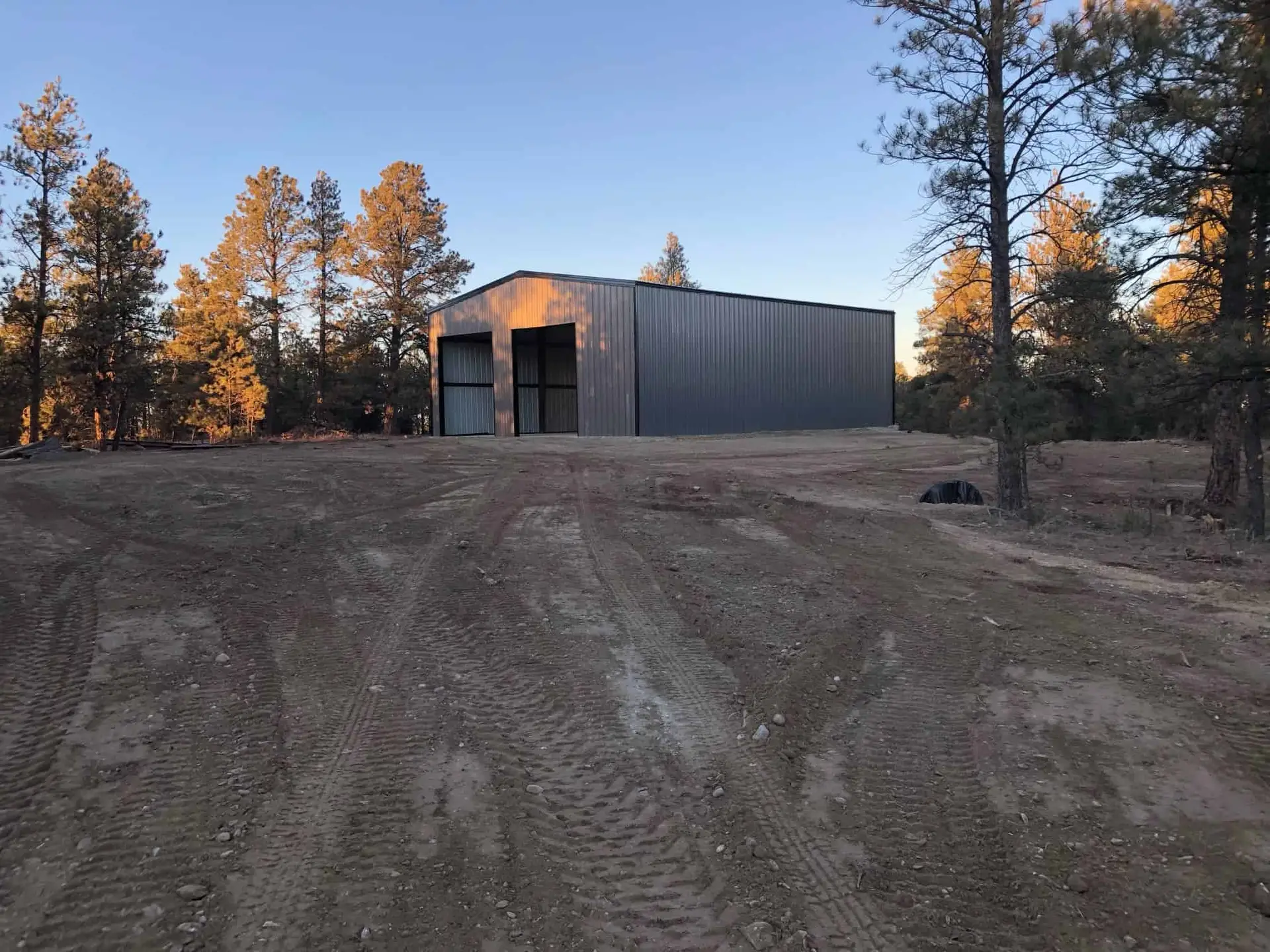 Gray metal-sided building with two large door openings on a freshly graded dirt site among ponderosa pines